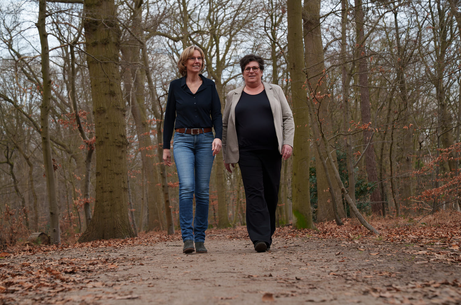 Twee personen maken een ontspannen wandeling in een bos, geschikt voor het bevorderen van welzijn en verbondenheid. Manuele therapie Prinsenbeek.